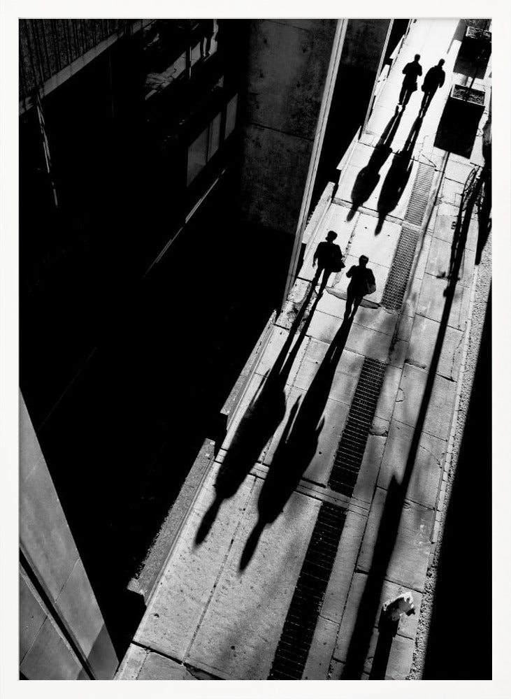 A dramatic high-angle, black and white photograph of several people walking on a sunlit sidewalk, casting long, stark shadows that stretch diagonally across the frame, contrasting with the deep black shadows of the adjacent building. Decor