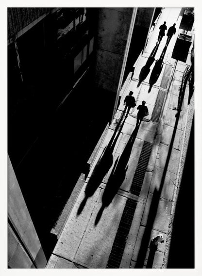 A dramatic high-angle, black and white photograph of several people walking on a sunlit sidewalk, casting long, stark shadows that stretch diagonally across the frame, contrasting with the deep black shadows of the adjacent building. Decor