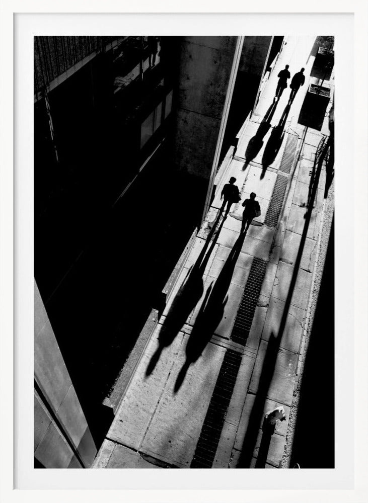 A dramatic high-angle, black and white photograph of several people walking on a sunlit sidewalk, casting long, stark shadows that stretch diagonally across the frame, contrasting with the deep black shadows of the adjacent building. Decor