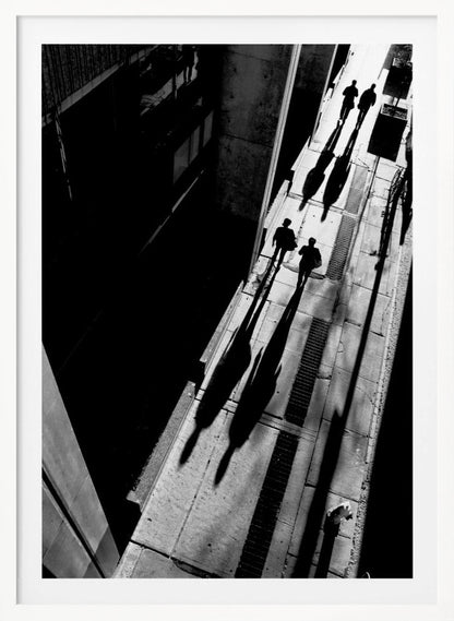 A dramatic high-angle, black and white photograph of several people walking on a sunlit sidewalk, casting long, stark shadows that stretch diagonally across the frame, contrasting with the deep black shadows of the adjacent building. Decor