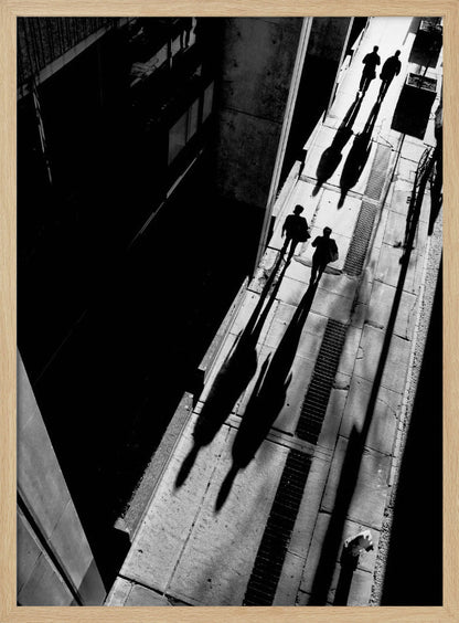 A dramatic high-angle, black and white photograph of several people walking on a sunlit sidewalk, casting long, stark shadows that stretch diagonally across the frame, contrasting with the deep black shadows of the adjacent building. Decor