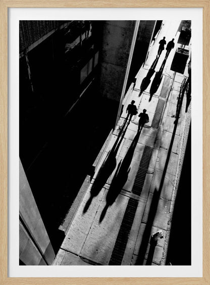 A dramatic high-angle, black and white photograph of several people walking on a sunlit sidewalk, casting long, stark shadows that stretch diagonally across the frame, contrasting with the deep black shadows of the adjacent building. Decor