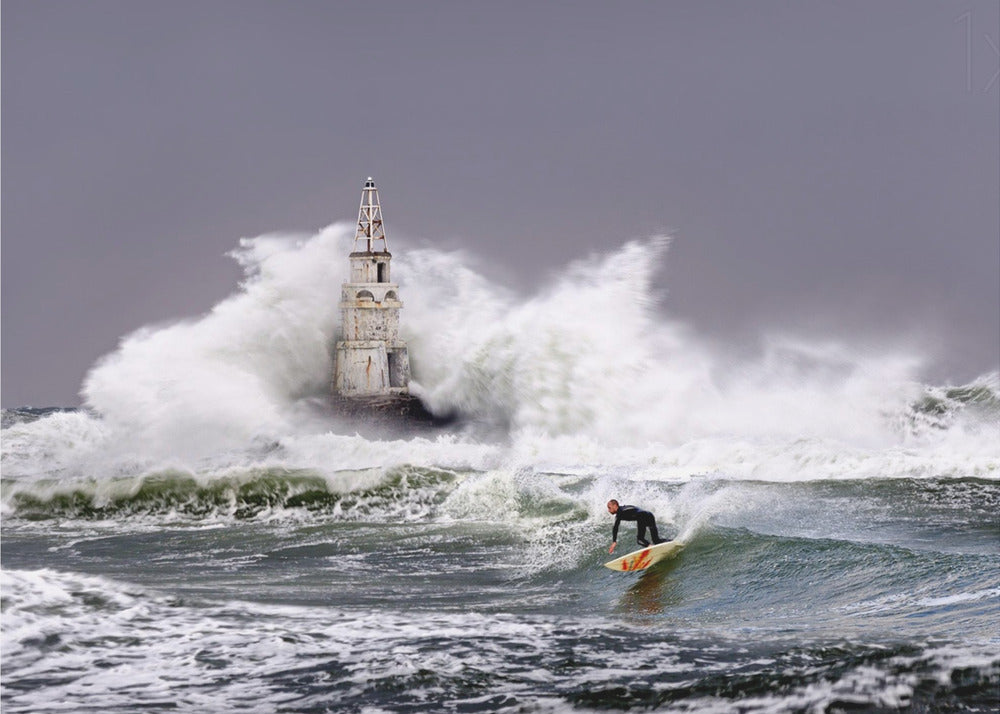 A framed photograph of a surfer in a black wetsuit riding a wave in a stormy sea. In the background, massive white waves crash against an old white lighthouse under a grey, overcast sky. Artwork