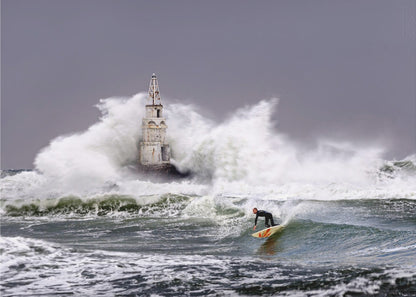 A framed photograph of a surfer in a black wetsuit riding a wave in a stormy sea. In the background, massive white waves crash against an old white lighthouse under a grey, overcast sky. Artwork