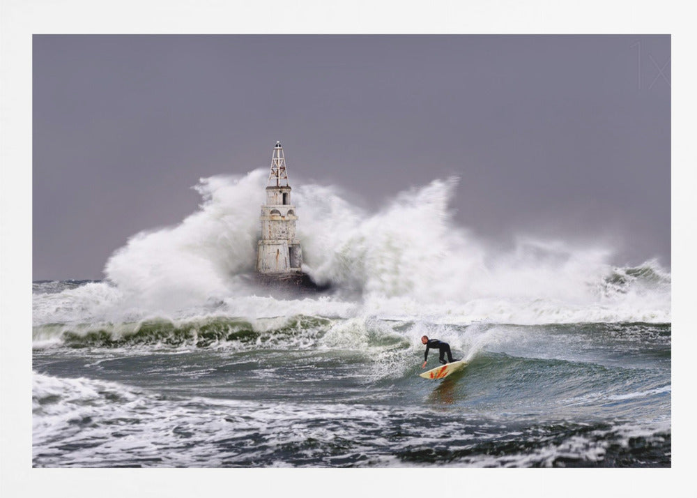 A framed photograph of a surfer in a black wetsuit riding a wave in a stormy sea. In the background, massive white waves crash against an old white lighthouse under a grey, overcast sky. Artwork