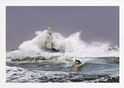 A framed photograph of a surfer in a black wetsuit riding a wave in a stormy sea. In the background, massive white waves crash against an old white lighthouse under a grey, overcast sky. Artwork