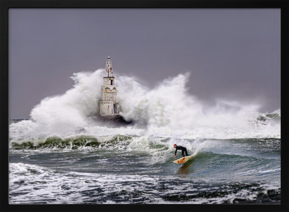 A framed photograph of a surfer in a black wetsuit riding a wave in a stormy sea. In the background, massive white waves crash against an old white lighthouse under a grey, overcast sky. Artwork