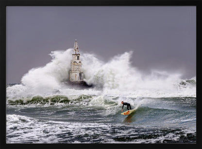 A framed photograph of a surfer in a black wetsuit riding a wave in a stormy sea. In the background, massive white waves crash against an old white lighthouse under a grey, overcast sky. Artwork