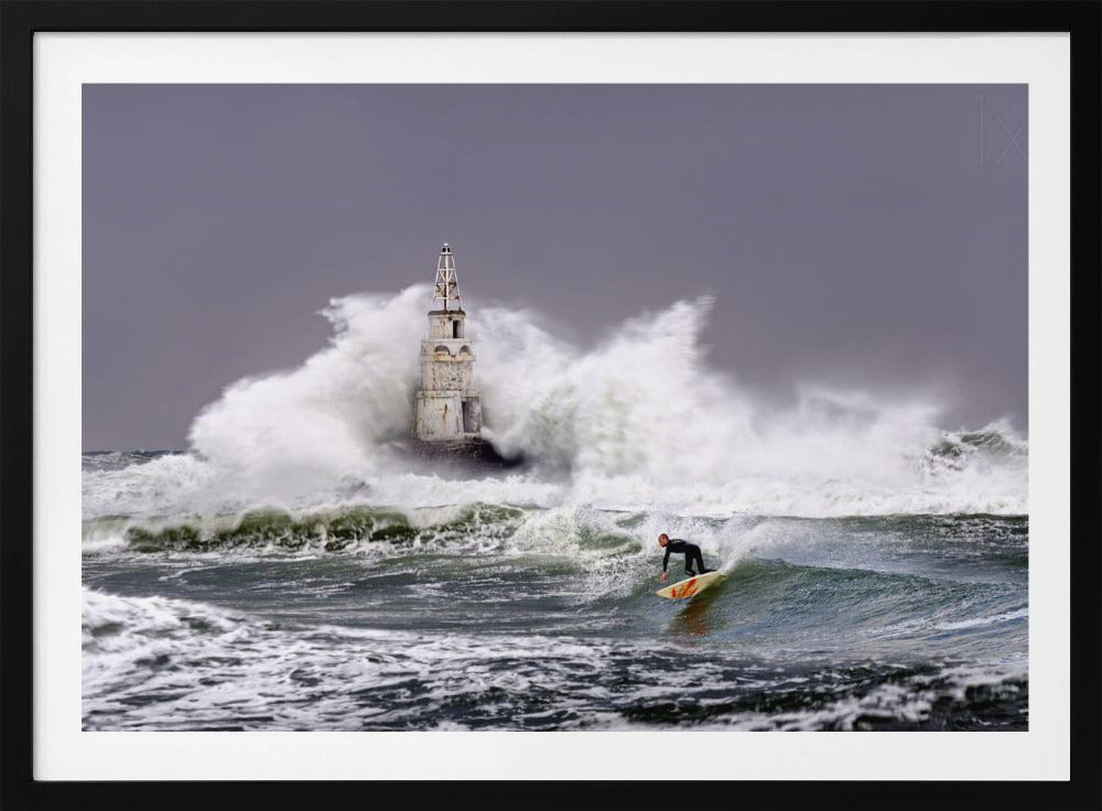 A framed photograph of a surfer in a black wetsuit riding a wave in a stormy sea. In the background, massive white waves crash against an old white lighthouse under a grey, overcast sky. Artwork
