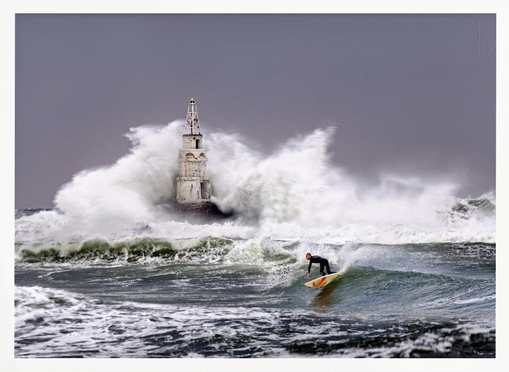A framed photograph of a surfer in a black wetsuit riding a wave in a stormy sea. In the background, massive white waves crash against an old white lighthouse under a grey, overcast sky. Artwork