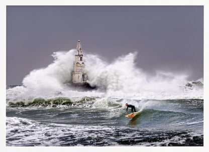 A framed photograph of a surfer in a black wetsuit riding a wave in a stormy sea. In the background, massive white waves crash against an old white lighthouse under a grey, overcast sky. Artwork