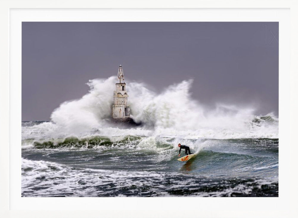 A framed photograph of a surfer in a black wetsuit riding a wave in a stormy sea. In the background, massive white waves crash against an old white lighthouse under a grey, overcast sky. Artwork