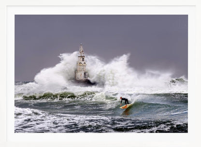 A framed photograph of a surfer in a black wetsuit riding a wave in a stormy sea. In the background, massive white waves crash against an old white lighthouse under a grey, overcast sky. Artwork