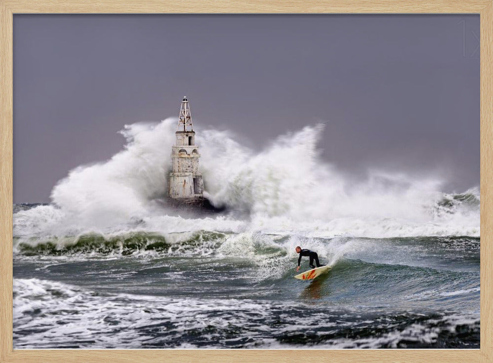 A framed photograph of a surfer in a black wetsuit riding a wave in a stormy sea. In the background, massive white waves crash against an old white lighthouse under a grey, overcast sky. Artwork