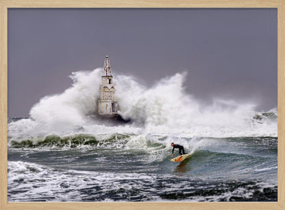 A framed photograph of a surfer in a black wetsuit riding a wave in a stormy sea. In the background, massive white waves crash against an old white lighthouse under a grey, overcast sky. Artwork