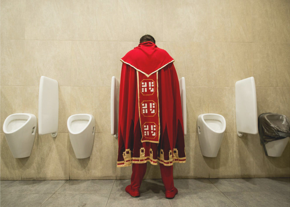 A back view of a person wearing an elaborate, long red and gold cape, standing at a urinal in a modern public restroom with beige tiled walls and multiple white urinals. Artwork