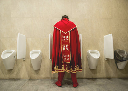 A back view of a person wearing an elaborate, long red and gold cape, standing at a urinal in a modern public restroom with beige tiled walls and multiple white urinals. Artwork