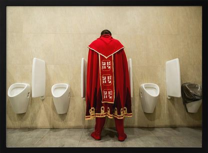 A back view of a person wearing an elaborate, long red and gold cape, standing at a urinal in a modern public restroom with beige tiled walls and multiple white urinals. Artwork