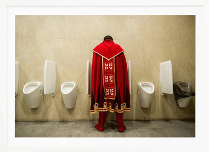 A back view of a person wearing an elaborate, long red and gold cape, standing at a urinal in a modern public restroom with beige tiled walls and multiple white urinals. Artwork