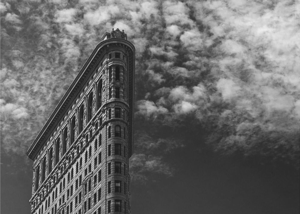 A dramatic low-angle black and white photograph of the Flatiron Building in New York City, with its sharp, triangular corner pointing towards a sky filled with fluffy, white clouds. The image is framed with a brushed silver border. Print