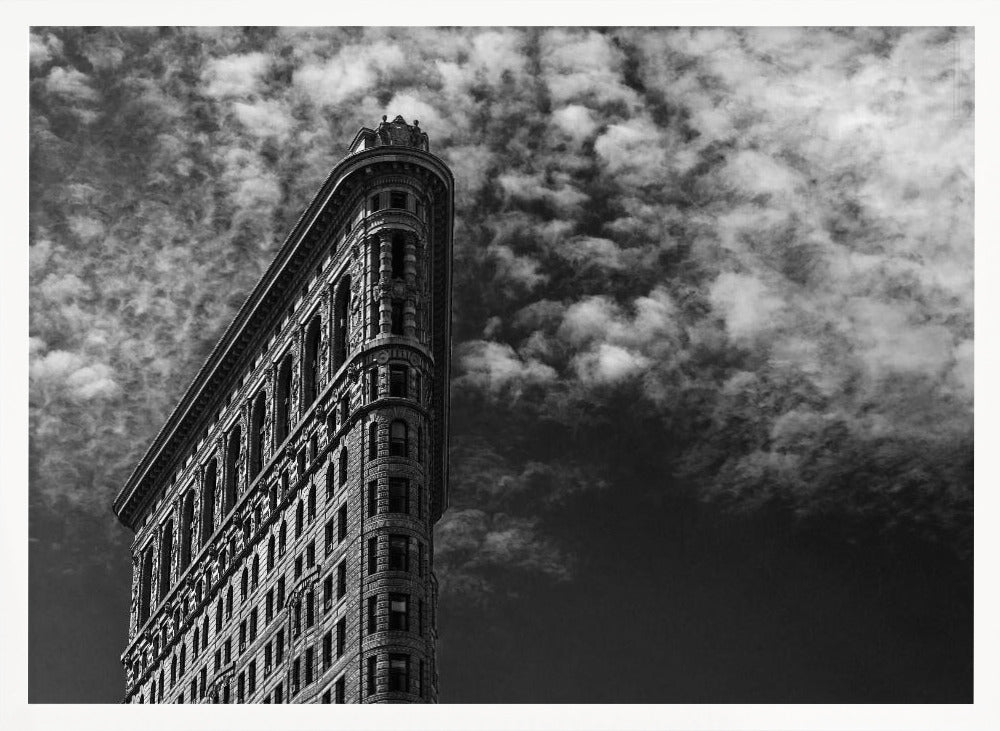 A dramatic low-angle black and white photograph of the Flatiron Building in New York City, with its sharp, triangular corner pointing towards a sky filled with fluffy, white clouds. The image is framed with a brushed silver border. Print