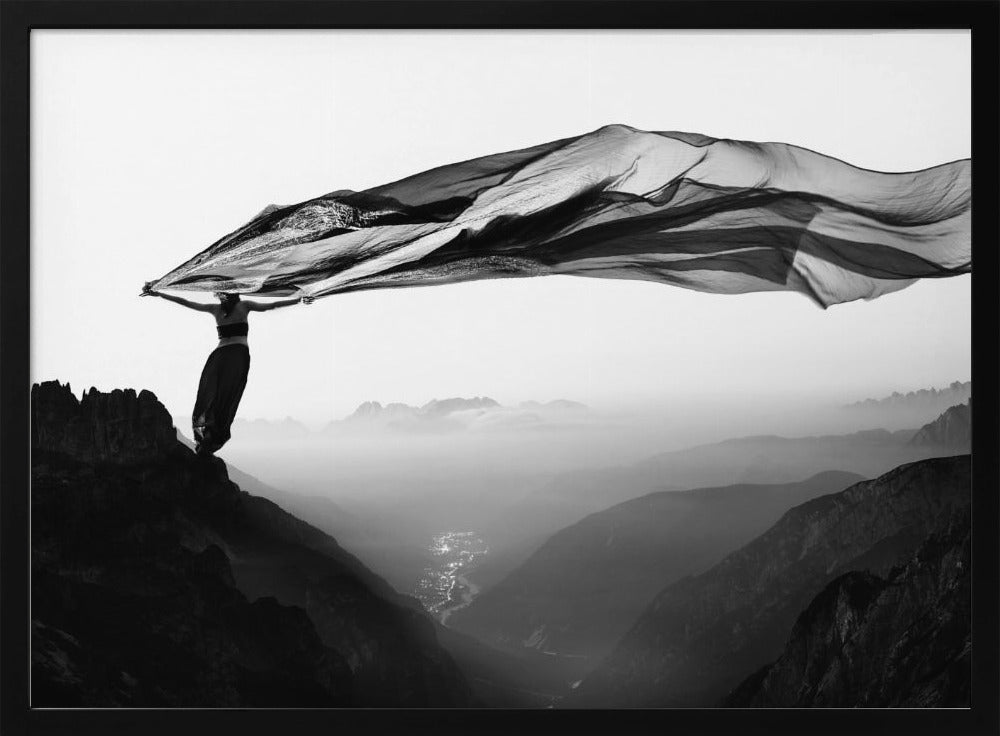 A dramatic black and white photograph of a woman standing on the edge of a mountain peak. She holds up a massive piece of dark, sheer fabric that billows in the wind, stretching across the sky. Below her, a vast, misty mountain range and a deep valley with city lights are visible. Wall Art