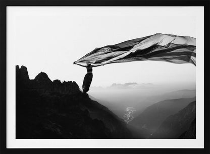 A dramatic black and white photograph of a woman standing on the edge of a mountain peak. She holds up a massive piece of dark, sheer fabric that billows in the wind, stretching across the sky. Below her, a vast, misty mountain range and a deep valley with city lights are visible. Wall Art