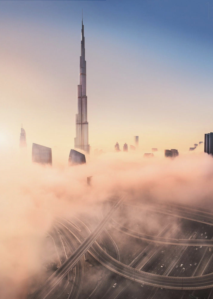 An aerial photograph of the Burj Khalifa in Dubai, towering over a thick blanket of pink and orange-hued fog at sunrise. Below the fog, a complex highway interchange is visible, with the clear blue sky above. Wall Art
