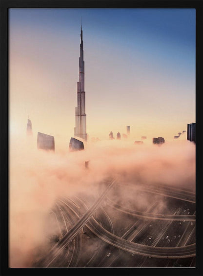 An aerial photograph of the Burj Khalifa in Dubai, towering over a thick blanket of pink and orange-hued fog at sunrise. Below the fog, a complex highway interchange is visible, with the clear blue sky above. Wall Art