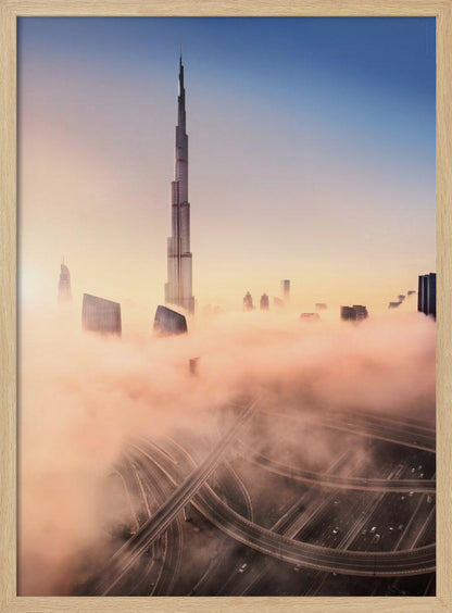 An aerial photograph of the Burj Khalifa in Dubai, towering over a thick blanket of pink and orange-hued fog at sunrise. Below the fog, a complex highway interchange is visible, with the clear blue sky above. Wall Art