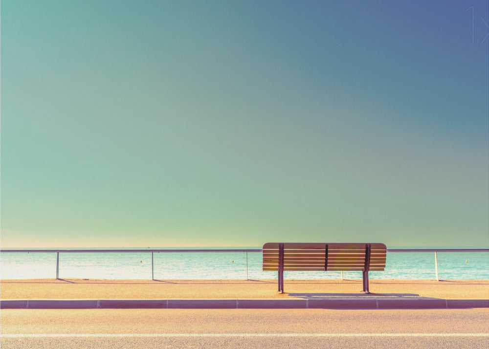 A minimalist, retro-toned photograph of a solitary wooden bench on a sunlit seaside promenade, overlooking a calm turquoise ocean under a vast, clear blue-green sky. Poster
