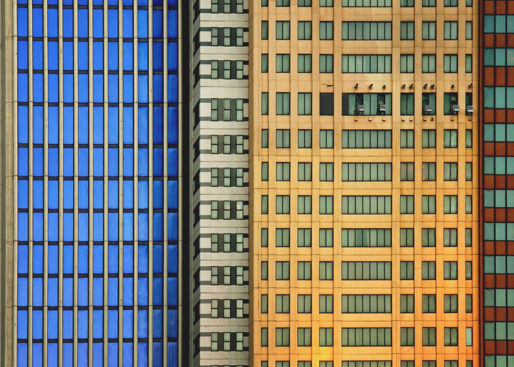 An abstract architectural photograph showing the colorful gridded facades of three adjacent skyscrapers, featuring a blue-windowed building, a white building, and a yellow building, all contained within a silver frame. Wall Art