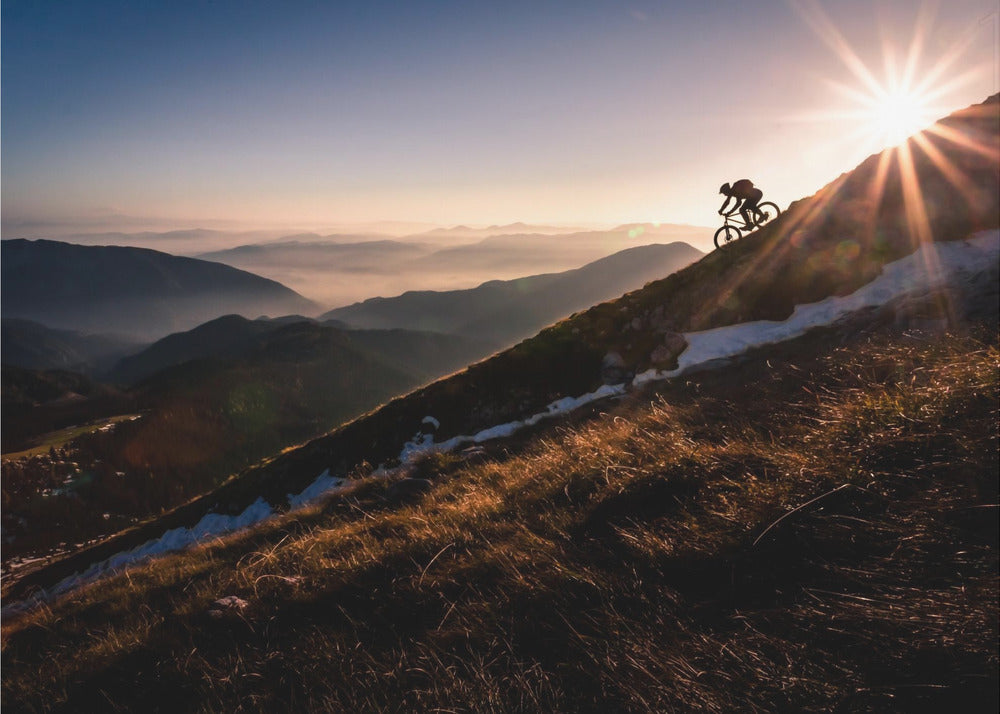 A silhouette of a mountain biker pushing their bike up a steep mountainside against a brilliant sunrise. The sun is cresting the peak, creating a starburst effect with long light rays. In the background, layers of mountains and valleys are filled with a soft morning mist under a clear gradient sky. Artwork