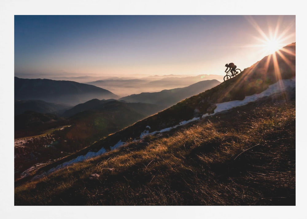 A silhouette of a mountain biker pushing their bike up a steep mountainside against a brilliant sunrise. The sun is cresting the peak, creating a starburst effect with long light rays. In the background, layers of mountains and valleys are filled with a soft morning mist under a clear gradient sky. Artwork