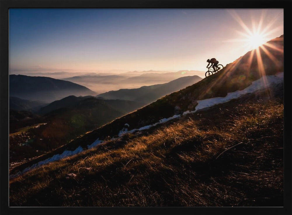 A silhouette of a mountain biker pushing their bike up a steep mountainside against a brilliant sunrise. The sun is cresting the peak, creating a starburst effect with long light rays. In the background, layers of mountains and valleys are filled with a soft morning mist under a clear gradient sky. Artwork