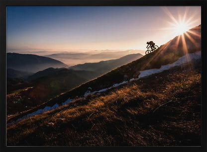 A silhouette of a mountain biker pushing their bike up a steep mountainside against a brilliant sunrise. The sun is cresting the peak, creating a starburst effect with long light rays. In the background, layers of mountains and valleys are filled with a soft morning mist under a clear gradient sky. Artwork