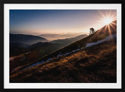 A silhouette of a mountain biker pushing their bike up a steep mountainside against a brilliant sunrise. The sun is cresting the peak, creating a starburst effect with long light rays. In the background, layers of mountains and valleys are filled with a soft morning mist under a clear gradient sky. Artwork