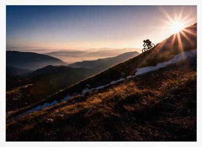 A silhouette of a mountain biker pushing their bike up a steep mountainside against a brilliant sunrise. The sun is cresting the peak, creating a starburst effect with long light rays. In the background, layers of mountains and valleys are filled with a soft morning mist under a clear gradient sky. Artwork