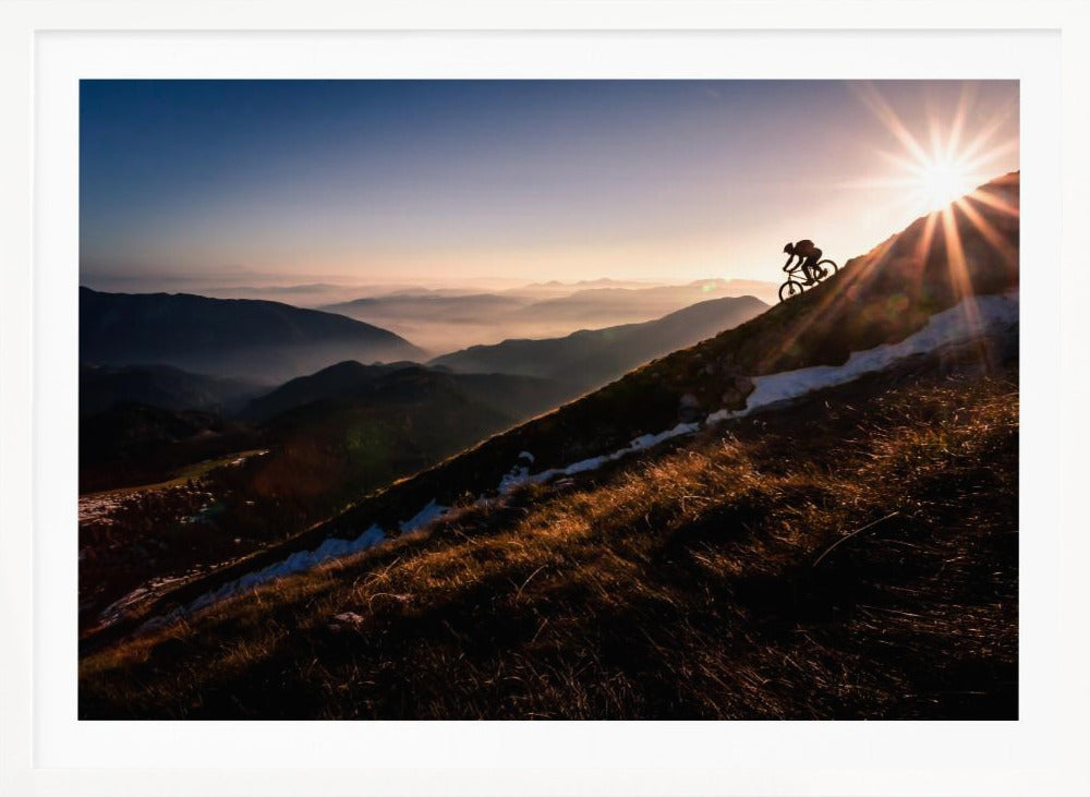 A silhouette of a mountain biker pushing their bike up a steep mountainside against a brilliant sunrise. The sun is cresting the peak, creating a starburst effect with long light rays. In the background, layers of mountains and valleys are filled with a soft morning mist under a clear gradient sky. Artwork