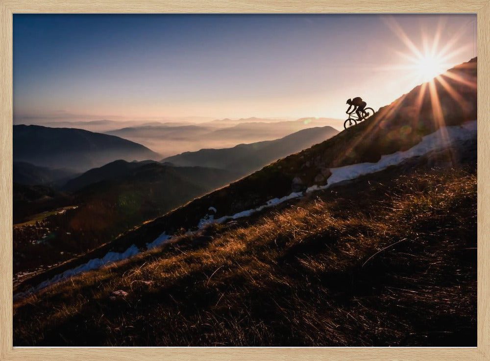A silhouette of a mountain biker pushing their bike up a steep mountainside against a brilliant sunrise. The sun is cresting the peak, creating a starburst effect with long light rays. In the background, layers of mountains and valleys are filled with a soft morning mist under a clear gradient sky. Artwork