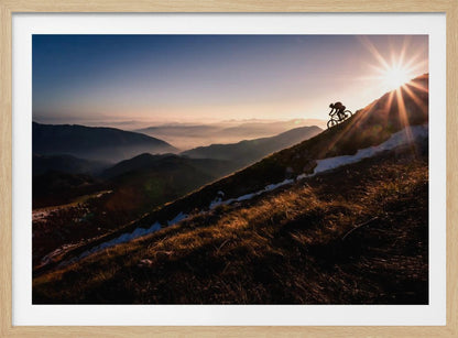 A silhouette of a mountain biker pushing their bike up a steep mountainside against a brilliant sunrise. The sun is cresting the peak, creating a starburst effect with long light rays. In the background, layers of mountains and valleys are filled with a soft morning mist under a clear gradient sky. Artwork
