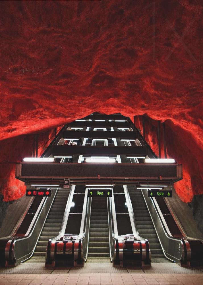 A dramatic low-angle shot looking up a series of escalators in a subway station with a cavernous, rough-textured ceiling glowing an intense, fiery red. Decor