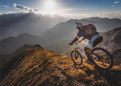 A mountain biker navigates a narrow, golden-lit ridge against a backdrop of a vast mountain range. The sun is breaking through clouds in the distance, casting dramatic rays of light across the epic landscape. Print