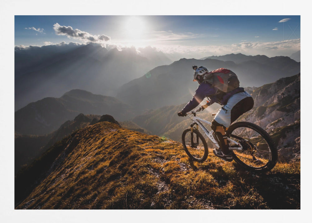 A mountain biker navigates a narrow, golden-lit ridge against a backdrop of a vast mountain range. The sun is breaking through clouds in the distance, casting dramatic rays of light across the epic landscape. Print