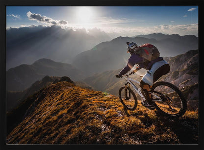 A mountain biker navigates a narrow, golden-lit ridge against a backdrop of a vast mountain range. The sun is breaking through clouds in the distance, casting dramatic rays of light across the epic landscape. Print
