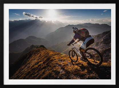 A mountain biker navigates a narrow, golden-lit ridge against a backdrop of a vast mountain range. The sun is breaking through clouds in the distance, casting dramatic rays of light across the epic landscape. Print