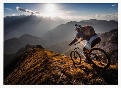 A mountain biker navigates a narrow, golden-lit ridge against a backdrop of a vast mountain range. The sun is breaking through clouds in the distance, casting dramatic rays of light across the epic landscape. Print