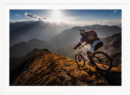 A mountain biker navigates a narrow, golden-lit ridge against a backdrop of a vast mountain range. The sun is breaking through clouds in the distance, casting dramatic rays of light across the epic landscape. Print