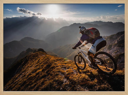 A mountain biker navigates a narrow, golden-lit ridge against a backdrop of a vast mountain range. The sun is breaking through clouds in the distance, casting dramatic rays of light across the epic landscape. Print