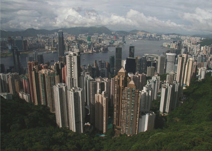 A high-angle photograph of the Hong Kong cityscape, viewed from a lush green peak. Numerous skyscrapers fill the frame, overlooking a bustling harbor with mountains in the distant background under a cloudy sky. The entire image is presented within a silver, brushed-metal frame. Artwork